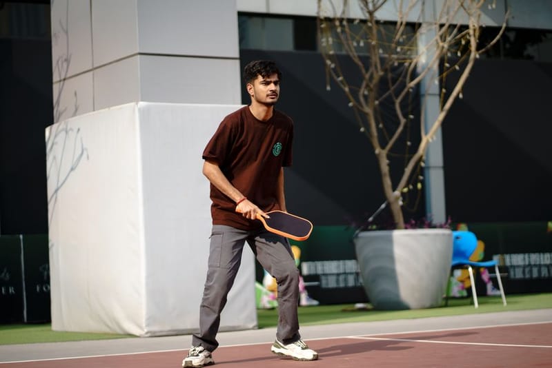 A man in a brown T-shirt holding a dark pickleball paddle with an orange edge on an outdoor court